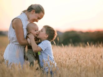 Mother holding baby and little boy kisses baby in wheat field